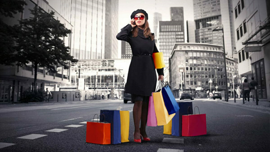 A woman holds her mobile phone to her ear, stood amid several brightly coloured bags of shopping