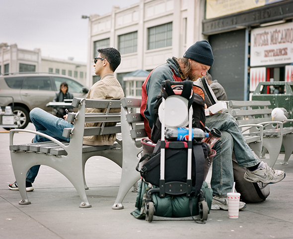 Two men sit on benches facing away from each other with their backs together, one seems comfortable and affluent whilst the other appears to have all their possessions in a case beside them