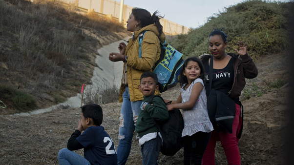 Children look frightened and upset as they and older relatives or companions are stopped at a border wall