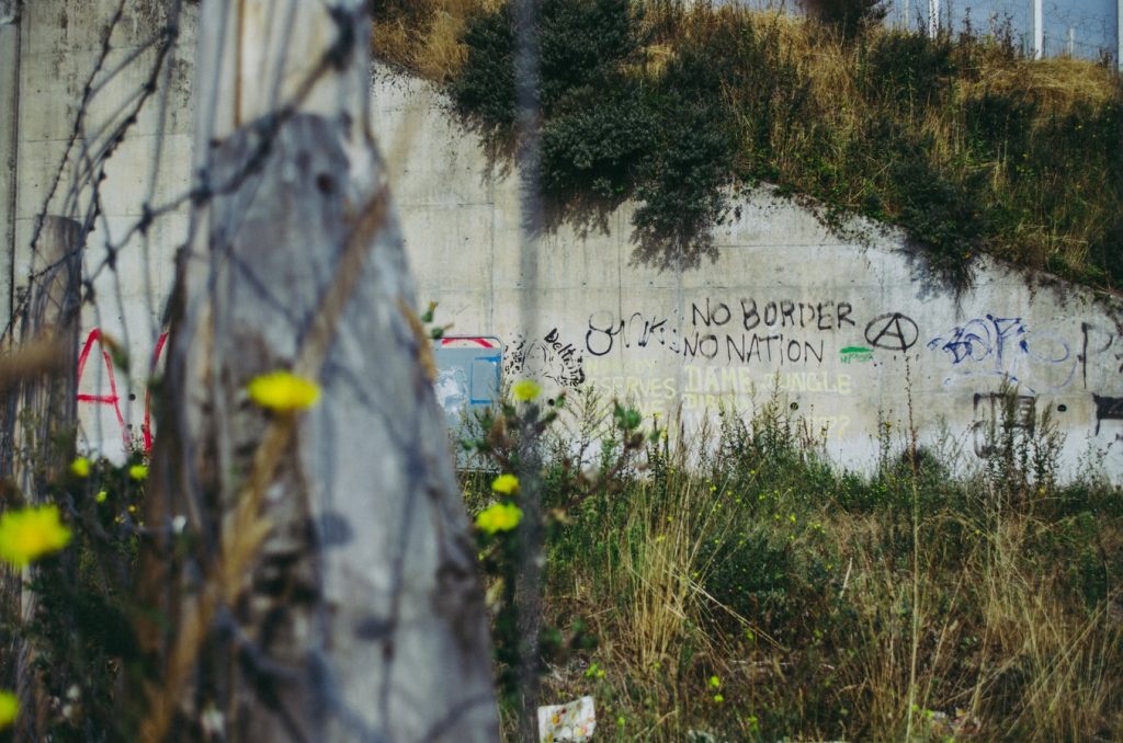 Graffiti reading 'No Borders no Nation' can be seen on exposed concrete amid weeds beneath a border fence