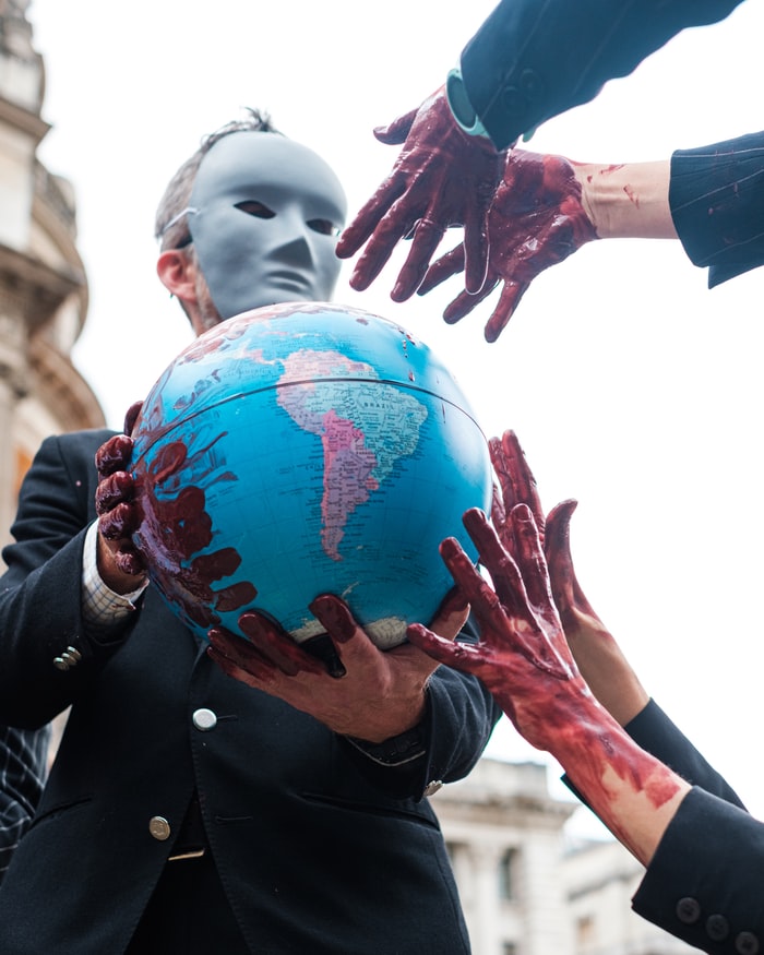 A masked man with red-painted hands (symbolising blood) clutches a globe, whilst other 'bloodied' hands reach towards it