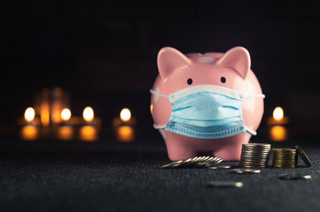 A piggy bank wearing a disposable mask stands in front of some blurry lights and behind stacks of coins