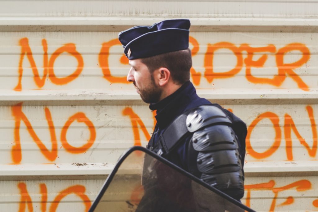 A man in a police uniform carrying a shield walks past graffiti appearing to say 'no borders'
