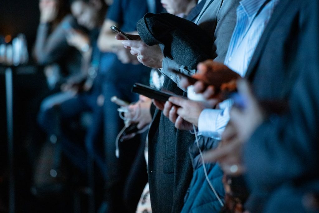 A row of mostly stood, mostly suited people looking down at their phones