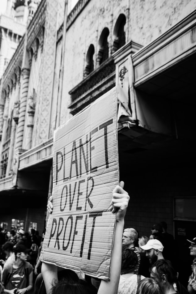 Black and white photograph of somebody holding a cardboard sign upon which has been written: 'PLANET OVER PROFIT'.