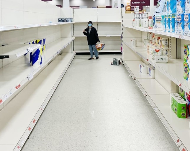 A woman wearing a mask stands at the end of a supermarket aisle where the shelves are almost bare