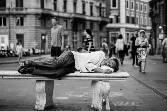 A homeless man sleeps on a bench in a busy city.