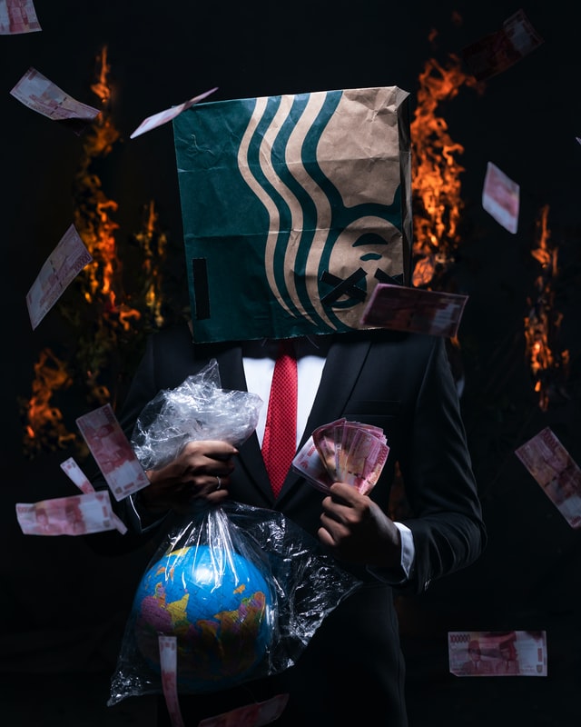 A man wearing various corporate symbols burns dollar bills and a model of the planet