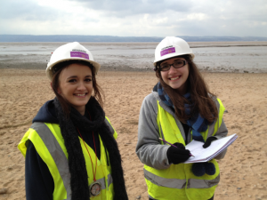 Two female students in high vis vest and protective hat smiling during fieldwork