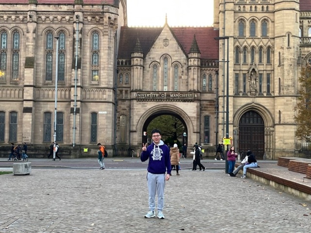A man doing a peace sign, standing in front of the University of Manchester arch