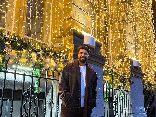Male student stood smiling in front of a building covered in lights