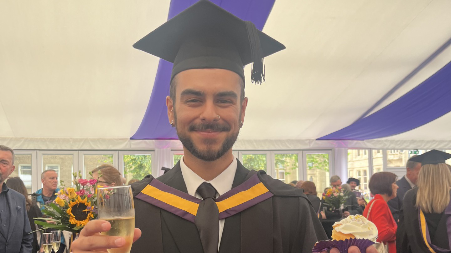 A man in a gazebo celebrates his graduation with a fizzy wine and a cupcake