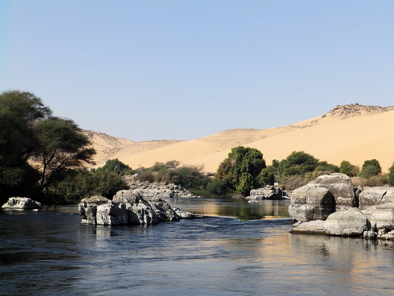 A small section of the river nile, featuring rocks and green trees around the river, with sandunes in the backdrop