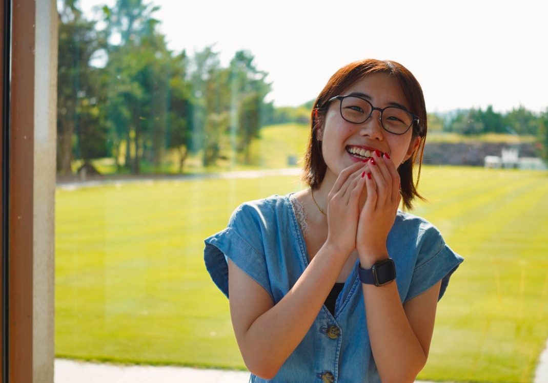AlumniPhoto-Akiko 800×560 Akiko stands in front of a window with a green garden behind her