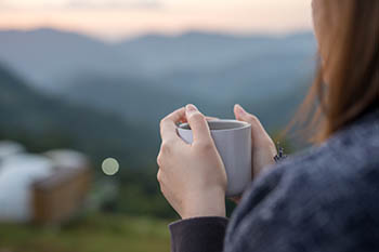 A person holding a mug of coffee, looking out over a hilly landscape.