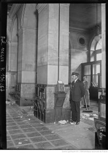 Photograph of two workers inside a colonnaded hall, one standing in uniform while another sweeps.
