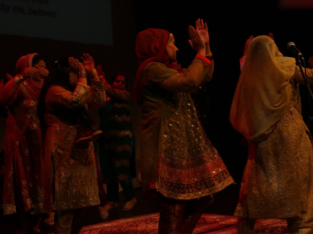 Ensemble of women reenacting a scene of traditional singing and dancing (Mehndi), common before the wedding day in South Asian culture. The Mehndi ceremony represents the bond of matrimony and signifies the love and affection between the couple and their families.