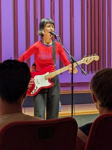 La Féline singer, Agnes, plays a bass guitar at the Lunch Time concert at the Martin Harris Centre. She is wearing a red top and black jeans and has her hair in a short black bob.