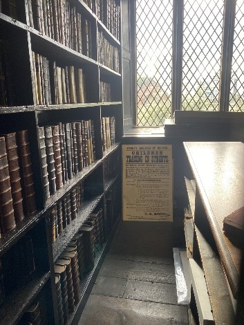 A view of a corner of the chambers. There is an old poster on the wall under the window - the headline reads CHILDREN TRADING IN THE STREETS but the remaining text is too small to read at this distance. A large old bookcase lines the wall to the left.